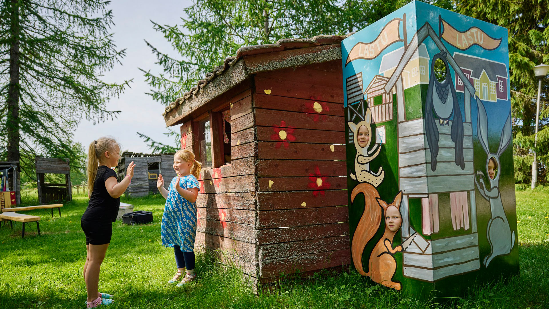 Lapsia majan vieressä ja majan sisällä. Kolme lasta kurkistaa majan aukoista ja kaksi leikkii ulkopuolella.