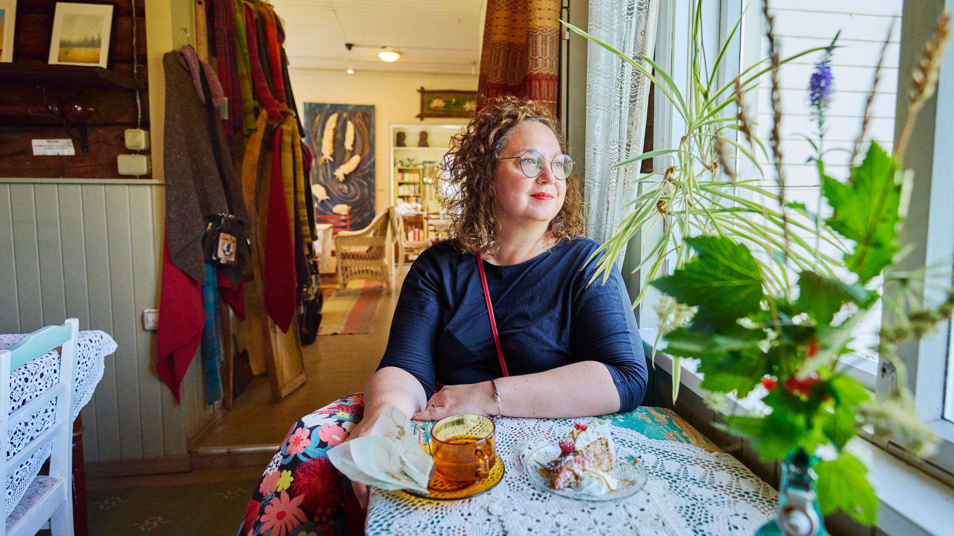 Nainen istuu pöydän ääressä edessään kahvikuppi ja kakkupala lautasella.