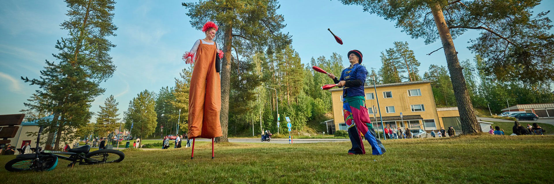 Kaksi sirkustaiteilijaa nurmikolla, joista toinen kävelee puujaloilla ja toinen jonglööraa keiloilla.