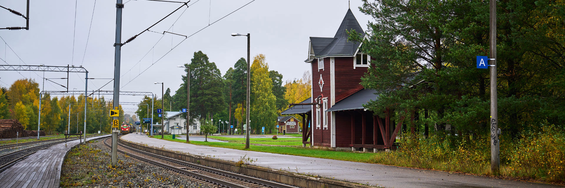 Junaradan raiteet, joiden päässä kaukaa näkyy saapuva juna. Asemalaiturin vieressä on vanha asema-alueen rakennus.